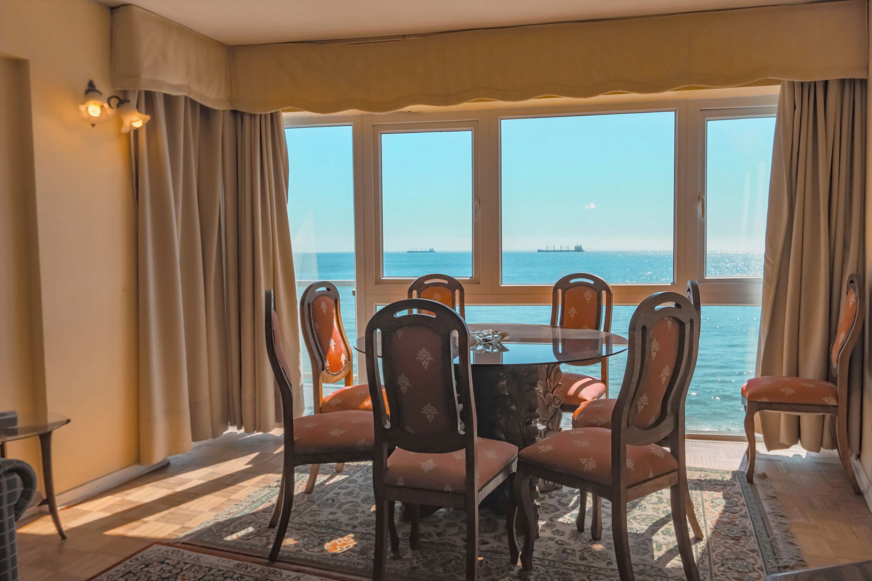 A dining area with a table and chairs, large windows with a view of the sea, and a patterned rug on the floor.