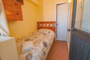 Een kleine kamer met een enkel bed, een houten hoofdbord, een nachtkastje en een deur met lichtschakelaar.