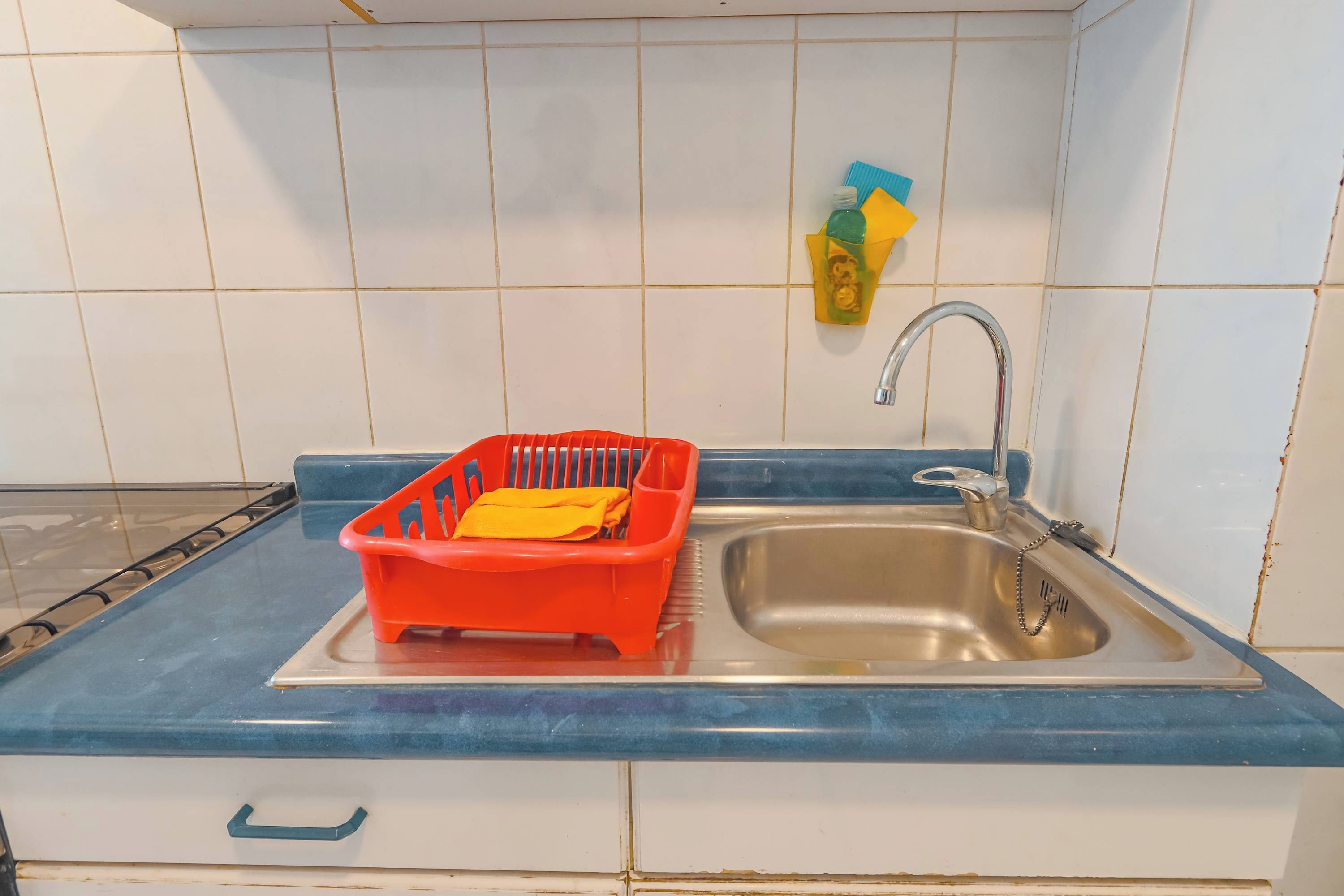 Cozinha com pia de aço inoxidável, escorredor de louça de plástico vermelho e bancada azul.