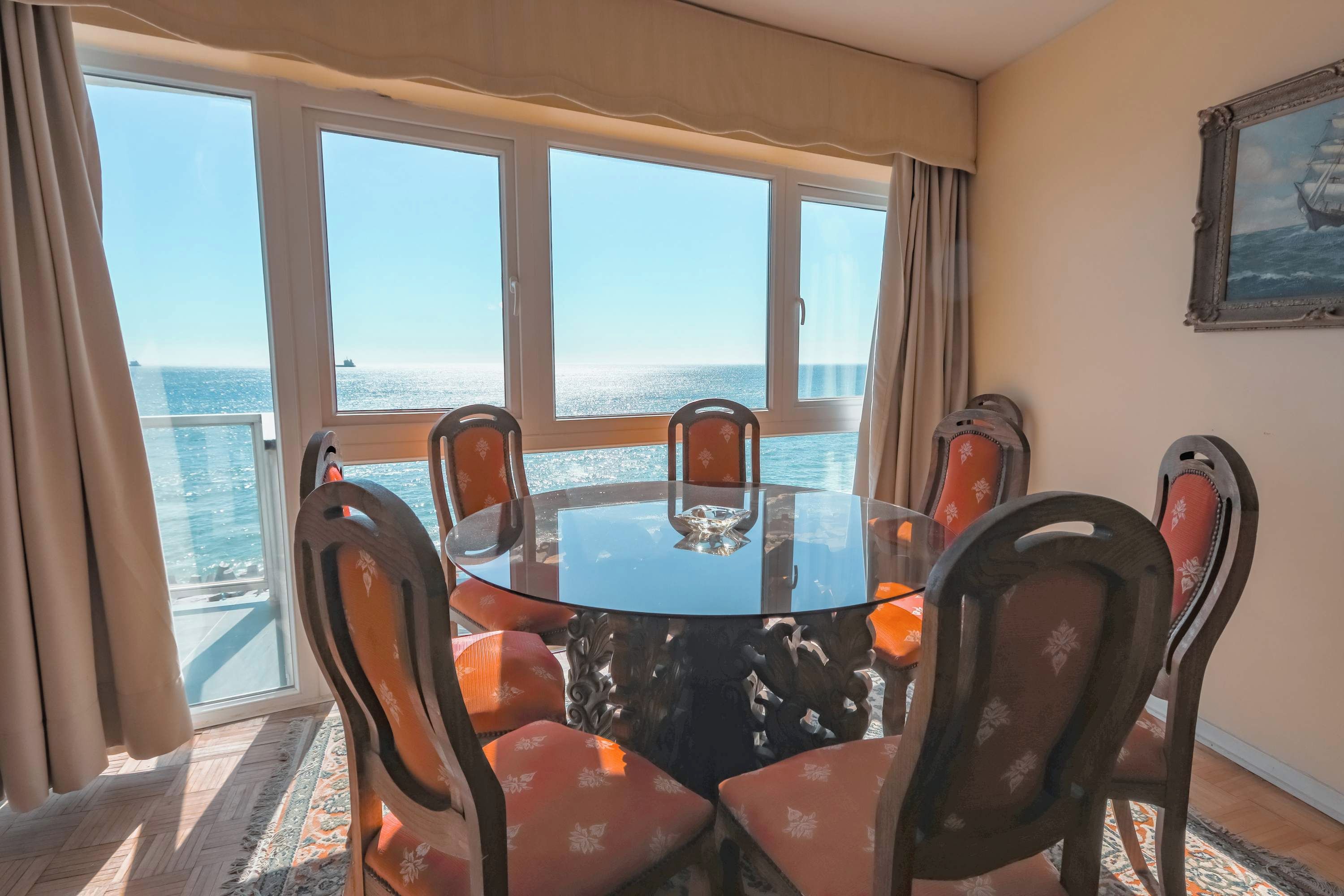 Una habitación con una mesa redonda de vidrio rodeada por seis sillas con tapicería floral, ventanas amplias con cortinas y un cuadro en la pared.