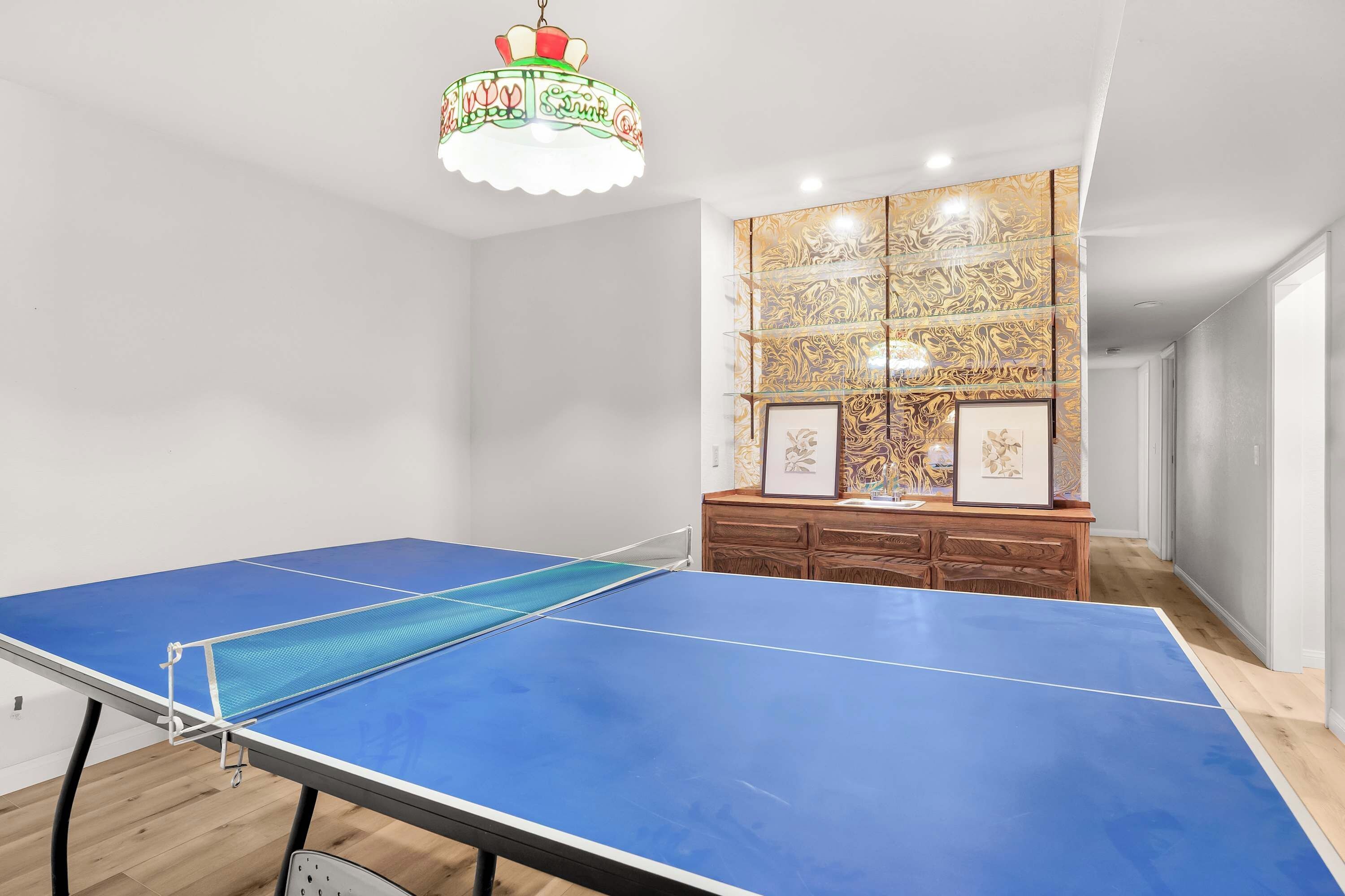 Una habitación con mesa de ping pong, un mueble de madera con marcos de fotos y un panel decorativo en la pared.