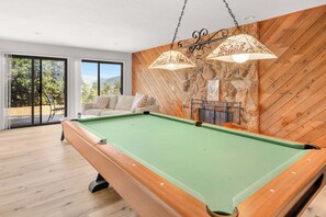Ein Billardtisch mit Holzrahmen und grüner Spielmatte, eine Couch mit gemusterten Kissen sowie eine Steinwand mit einem angebrachtem Schild.