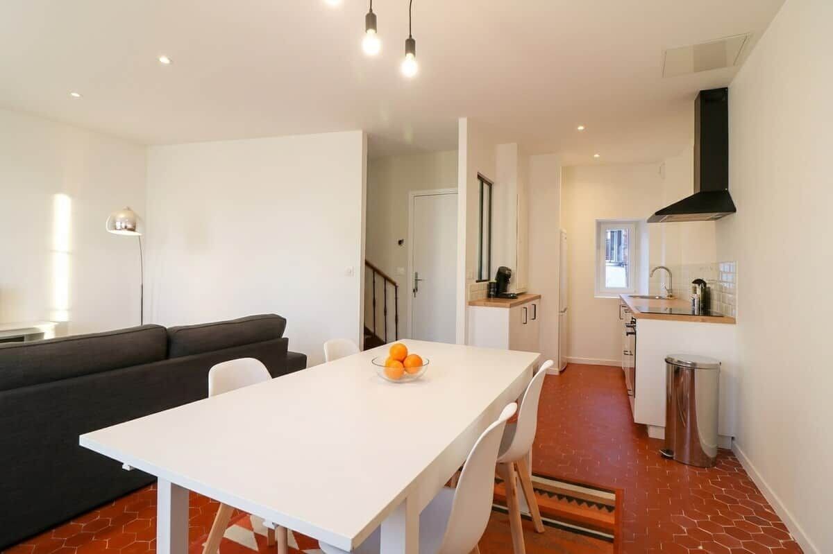 Une cuisine moderne avec un plan de travail blanc, une hotte noire et un coin repas comprenant une table et des chaises blanches.