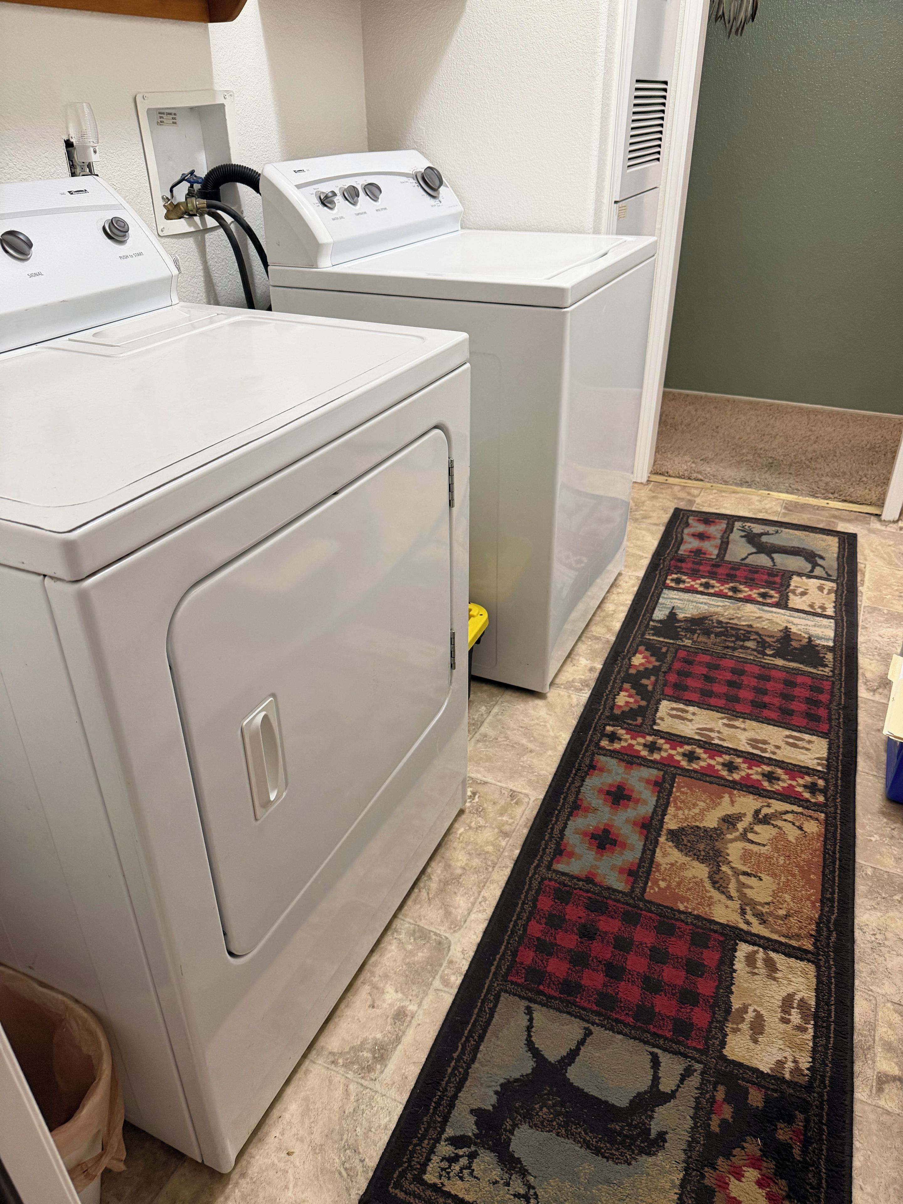 A laundry room with two white front-loading washers and dryers, a patterned rug, and a vent on the wall.