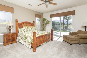 A bedroom with a wooden bed, a nightstand, a lamp, a sofa, and a view of the outdoors through a sliding glass door. - 5 Star Rating Oasis Golf Pool Spa 3BED Pga West (La Quinta)