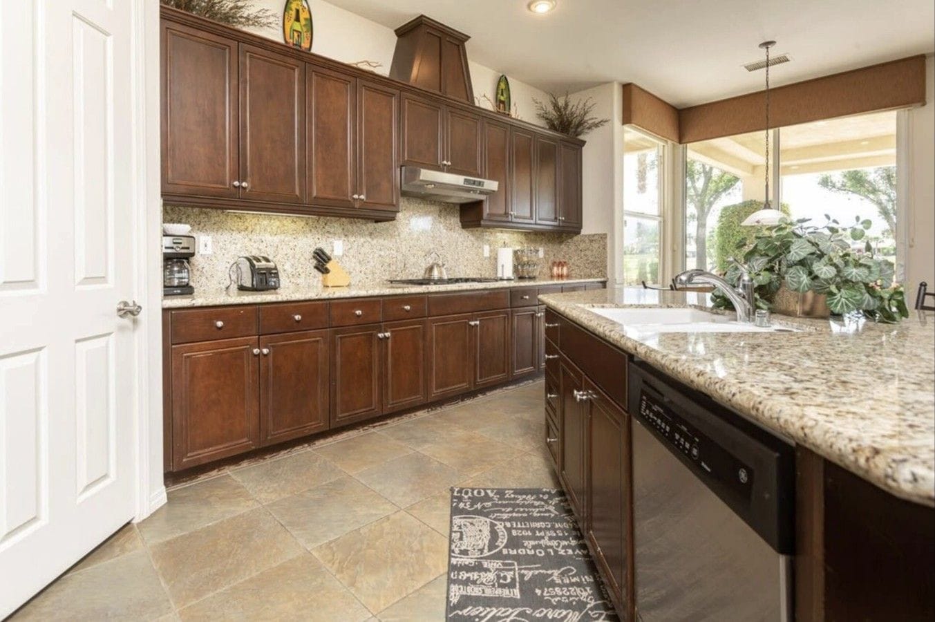 A kitchen with dark wooden cabinets, granite countertops, a built-in dishwasher, and a window providing natural light.
