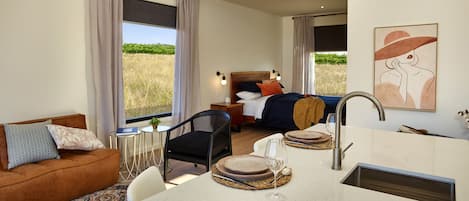 A hotel room with a bed, a dining area with a table and chairs, a sofa, and a large window with a view of a field.