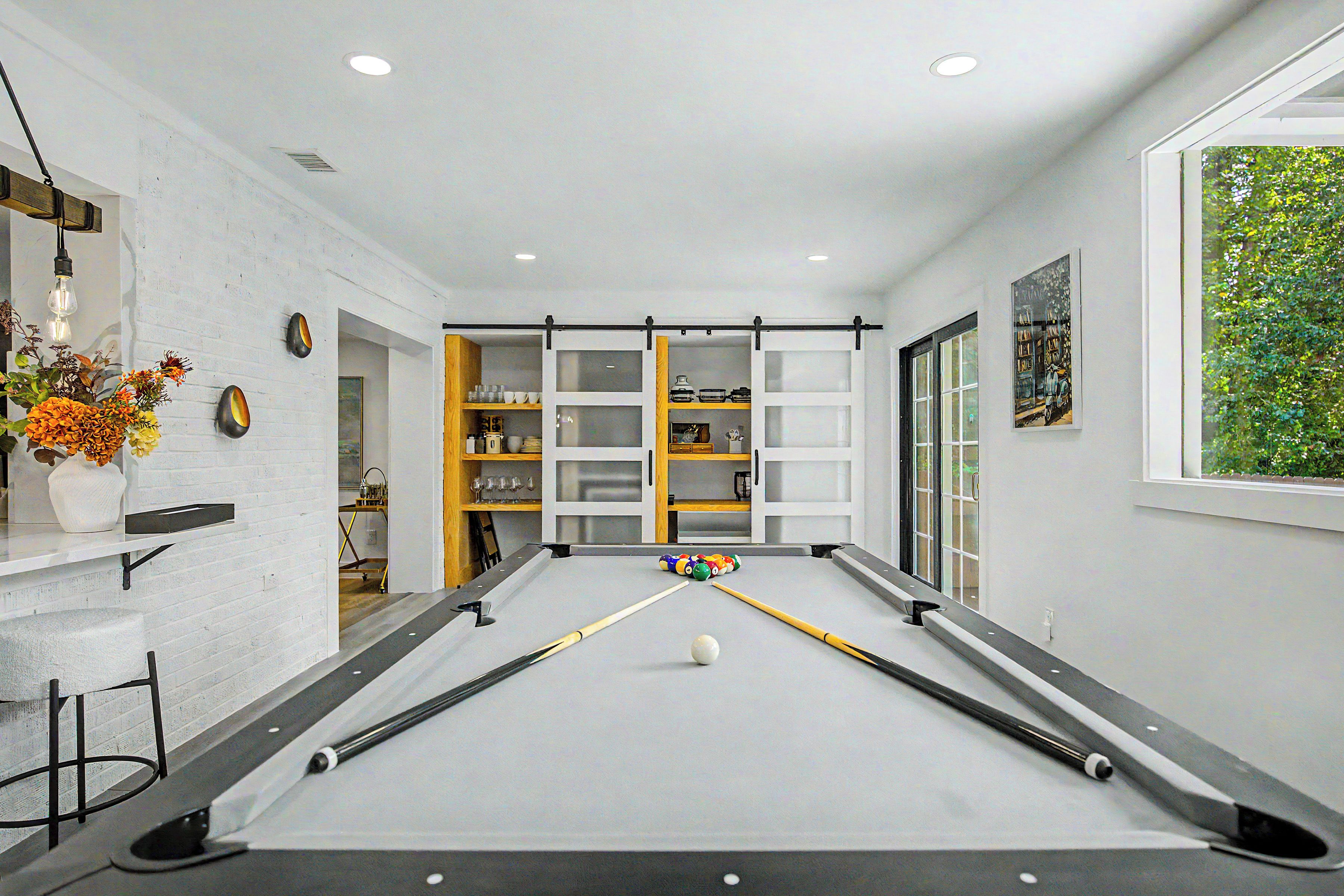 A pool table with a billiard cue, a vase with flowers, and a window with a view of greenery.