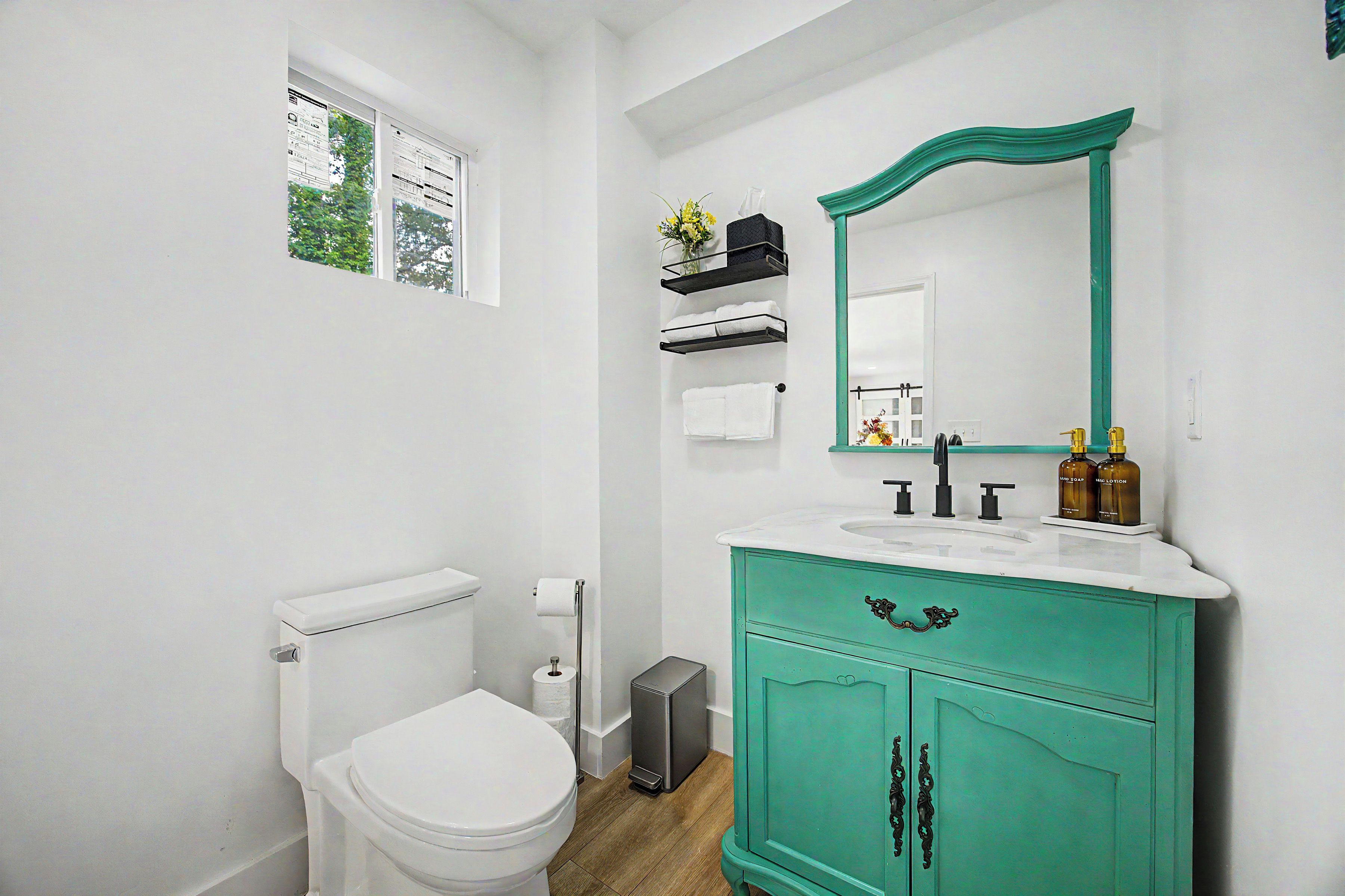 A bathroom with a white toilet, a green vanity with a mirror, and a window with a view of trees.