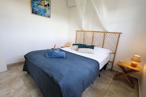 Ein ordentlich bezogenes Bett mit blauer Bettdecke, ein aufgerolltes Handtuch und eine dekorative, orangefarbene Blume. Das hölzerne Kopfteil weist ein geometrisches Muster auf, ein Nachttisch mit Lampe und ein Bild an der Wand sind ebenfalls zu sehen.