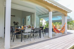 Een overdekte eethoek met tafels en stoelen, een hangmat en uitzicht op de tuin.