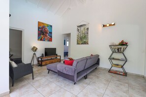 Ein Wohnzimmer mit grauem Sofa, einem Fernseher auf einem Holzständer, einer Stehlampe und einem Bücherregal mit Büchern und Blumen.
