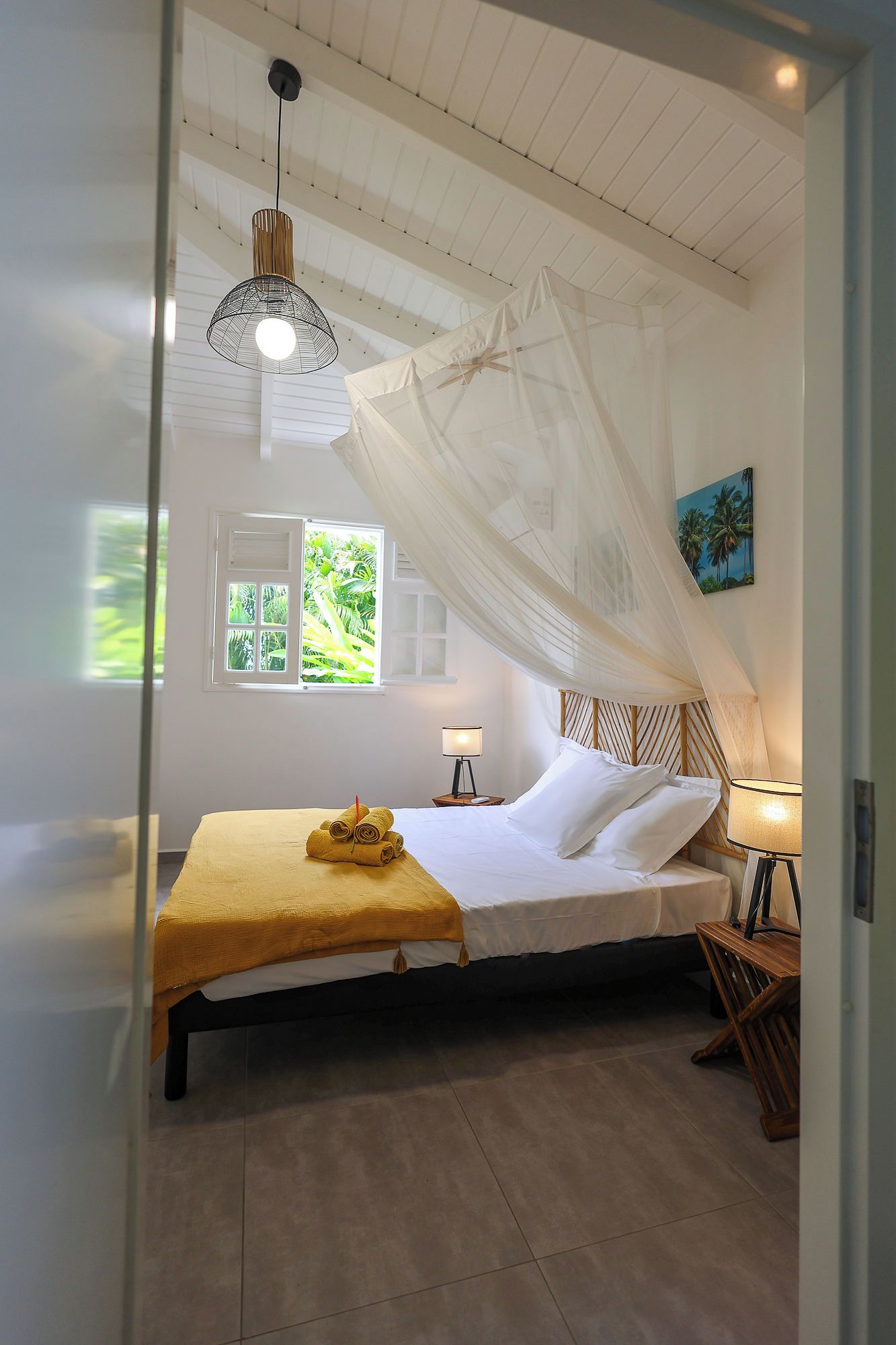 A bedroom with a bed, a bedside table, a lamp, a window with a view of greenery, and a ceiling fan.