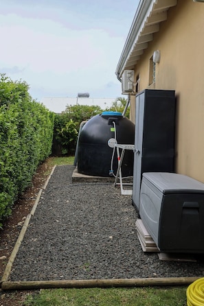 Een buitenruimte met een waterreservoir, een grijs kastje en een houten omheining.