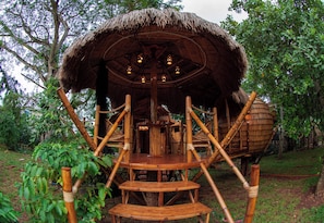 A thatched-roof structure with a wooden staircase leading to an upper level, surrounded by greenery.