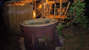 Une construction rustique à toit de chaume, dotée d’un intérieur en bois, entourée de murs en bambou et agrémentée d’une grande fontaine en bois.