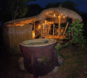 Une construction rustique à toit de chaume, dotée d’un intérieur en bois, entourée de murs en bambou et agrémentée d’une grande fontaine en bois.