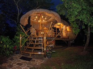 A thatched-roof structure with a circular central area, surrounded by a bamboo framework, and a stone-paved entrance.
