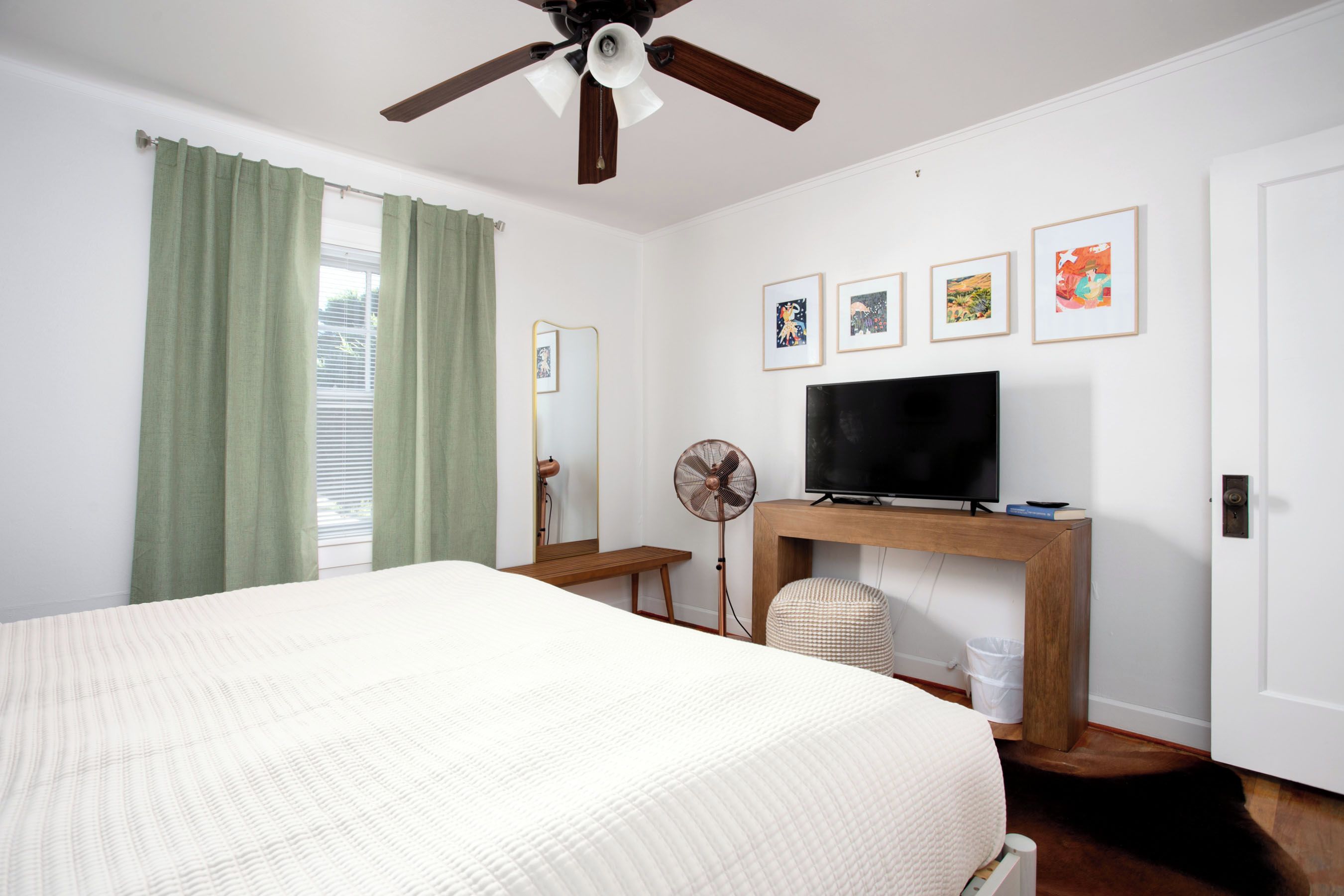 Quarto com cama, ventoinha no teto, TV sobre um suporte de madeira e arte na parede.