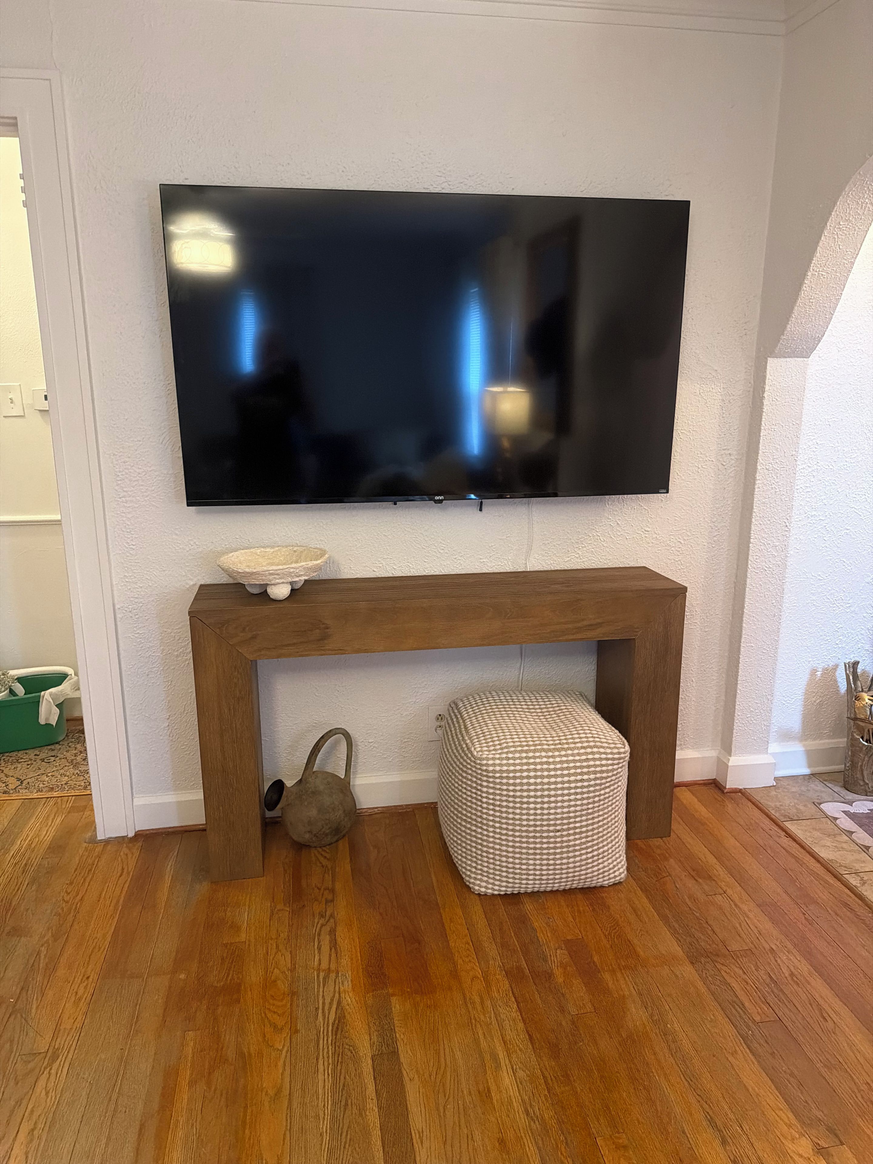 A flat-screen TV mounted on a wall above a wooden console table with a woven basket and a small stool.