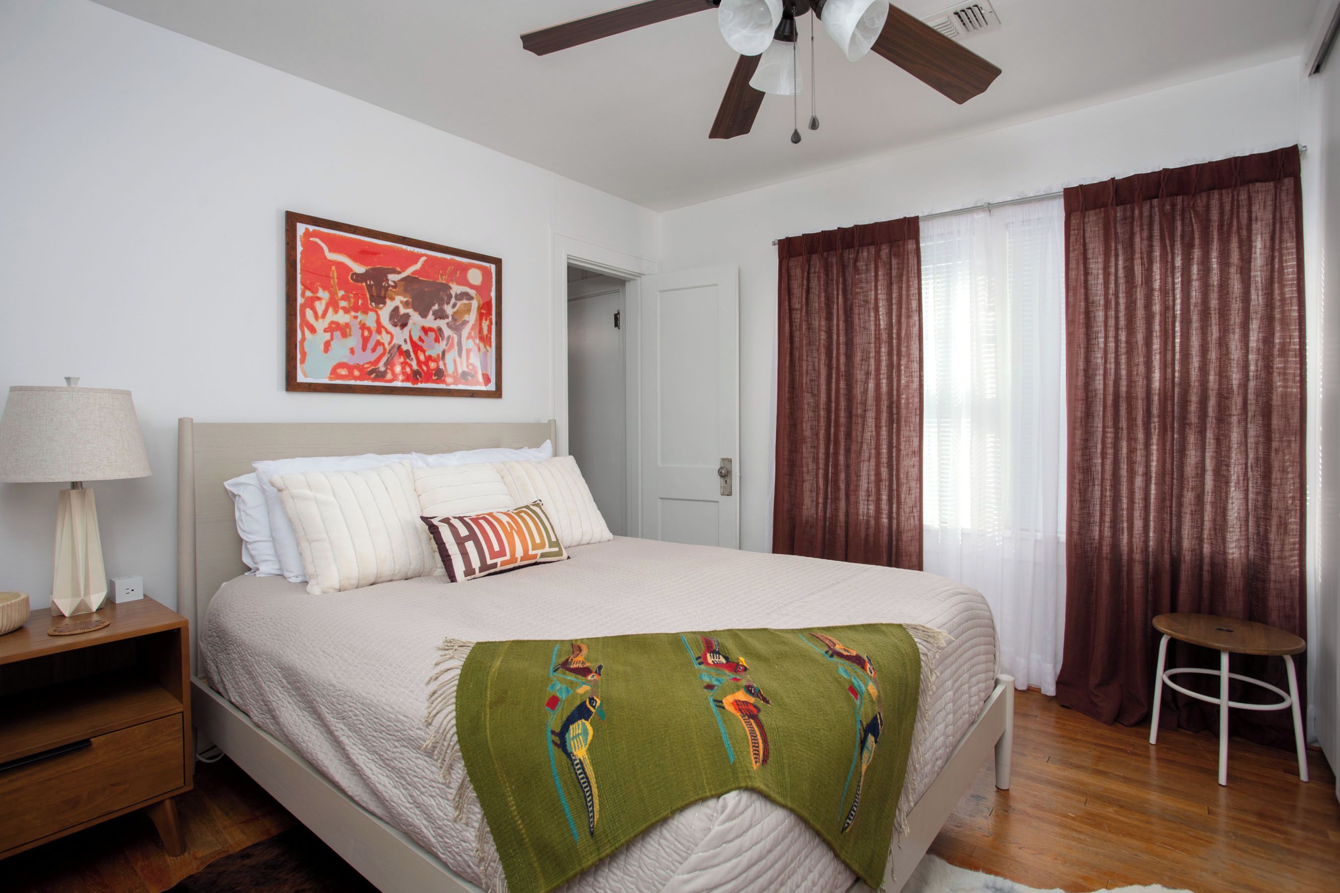 A bedroom with a bed, a nightstand, a lamp, a ceiling fan, a door, and a window with curtains.