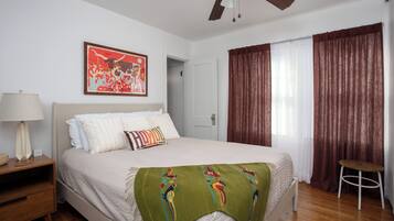 A bedroom with a bed, a nightstand, a lamp, a ceiling fan, a door, and a window with curtains.