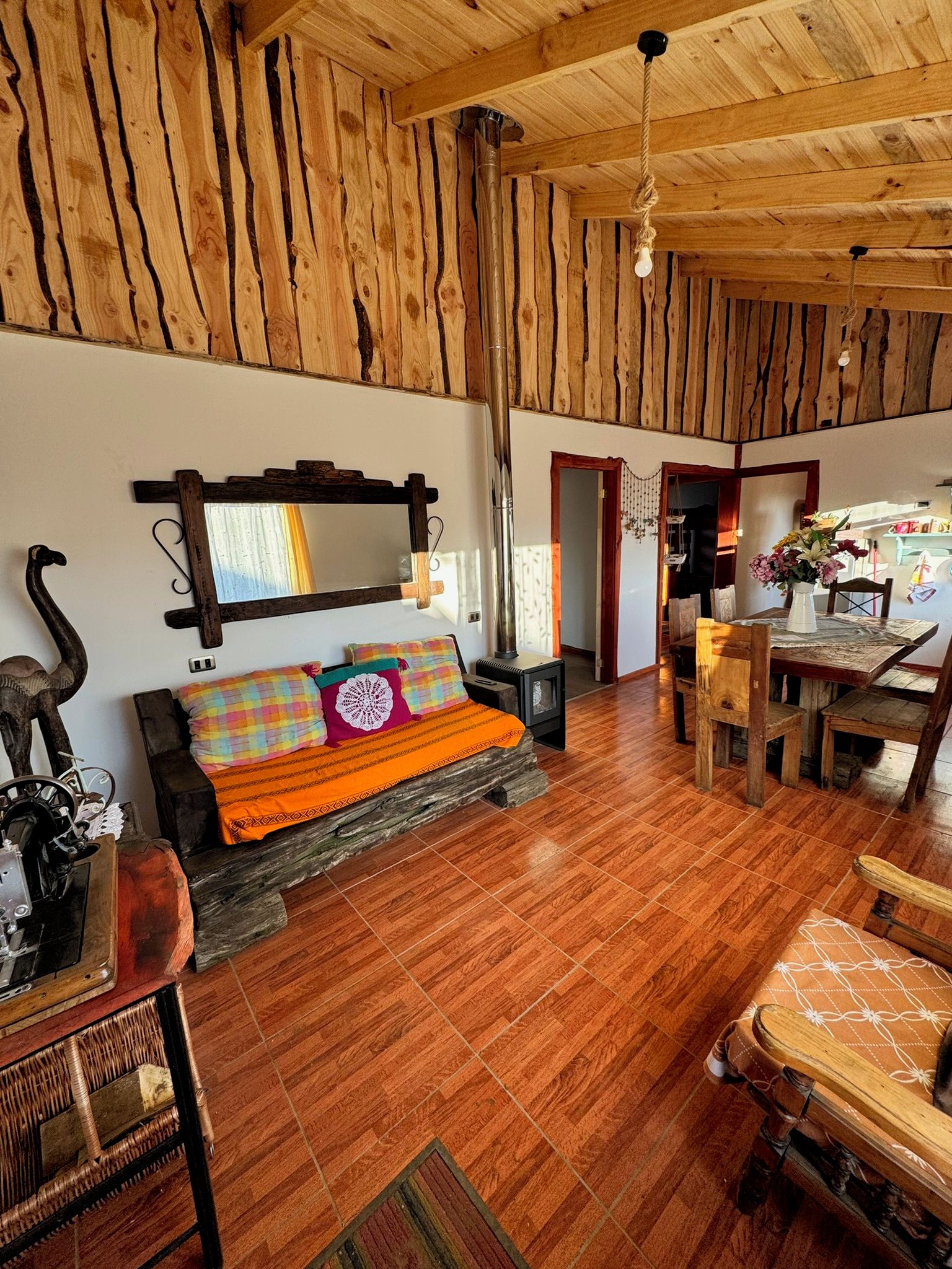 A rustic interior with a wooden ceiling, a sofa with colorful cushions, a dining table, and a chair.