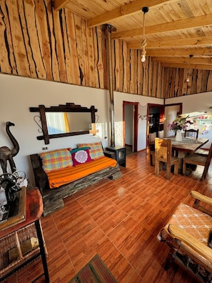 A rustic interior with a wooden ceiling, a sofa with colorful cushions, a dining table, and a chair.