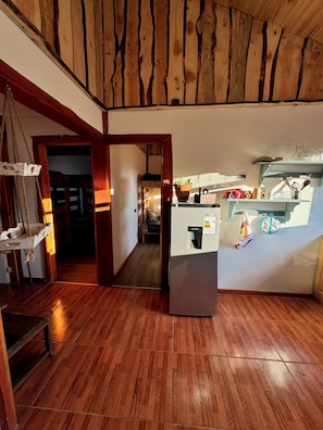 A room with a wooden floor, a kitchen area with a refrigerator, and a doorway leading to another room.