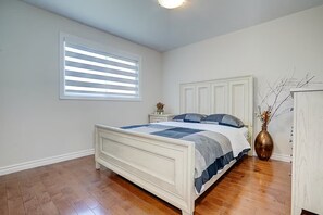 Ein Schlafzimmer mit einem Bett, einem Nachttisch, einer Kommode, einem Fenster mit Jalousien und einer Vase mit Zweigen.