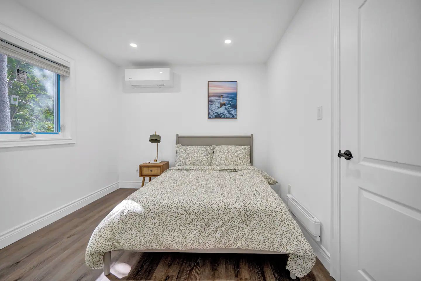 A bedroom with a bed, a nightstand, a window, an air conditioning unit, and a door.