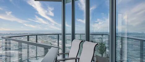 A high-rise balcony with a clear view of the cityscape, a glass railing, and a single chair.