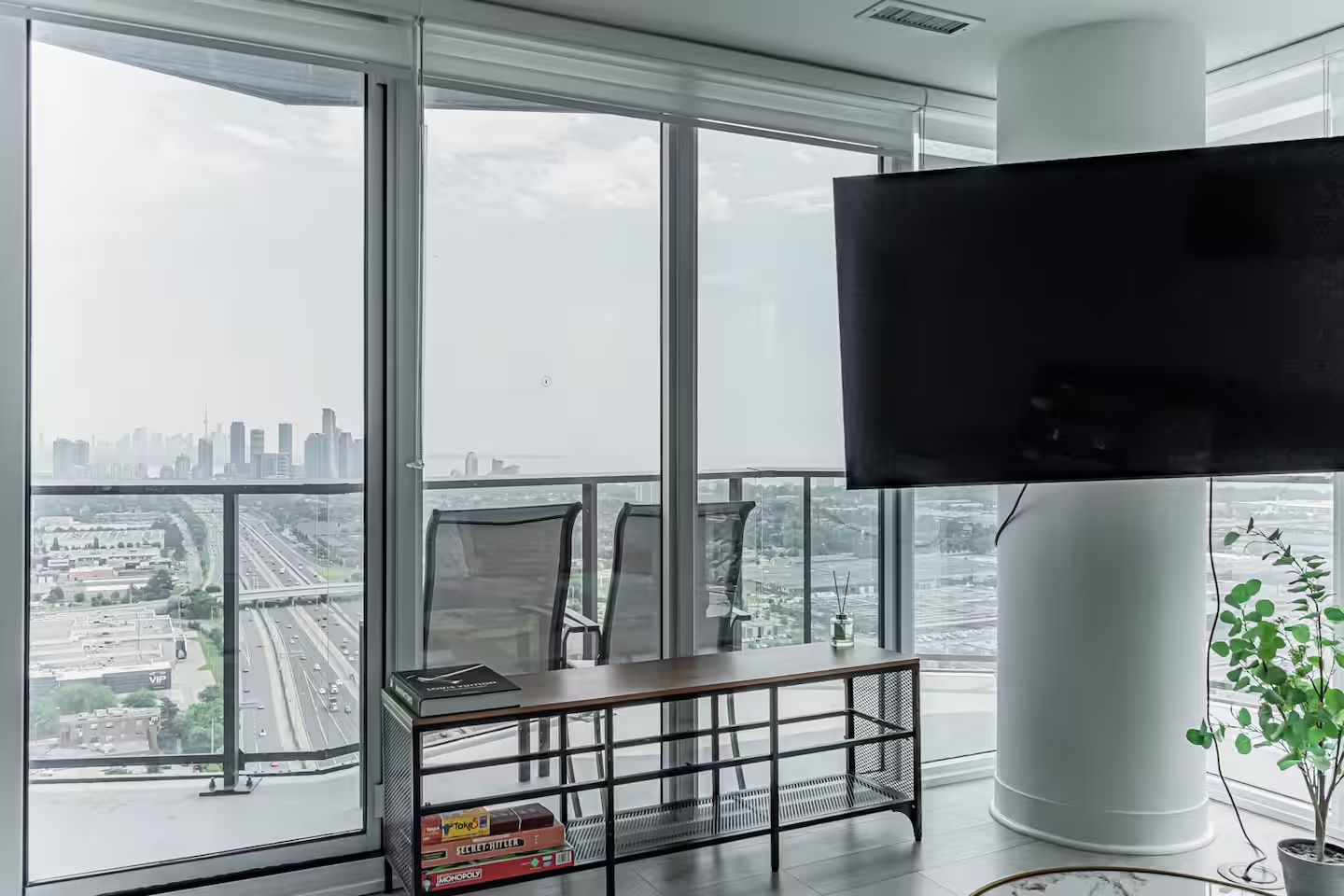 Un salon moderne doté d’un grand téléviseur, d’une bibliothèque remplie de jeux de société et offrant une vue sur le paysage urbain grâce à de grandes fenêtres du sol au plafond.