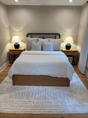 A neatly made bed with a white duvet, flanked by two bedside tables with lamps, in a room with wooden flooring and a white rug. - A frame cabin (Swannanoa)