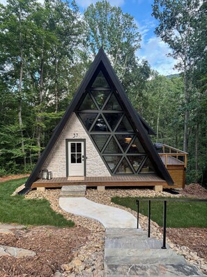 A modern, triangular-shaped cabin with a stone facade, a wooden deck, and a clear view of the surrounding forest. - A frame cabin (Swannanoa)