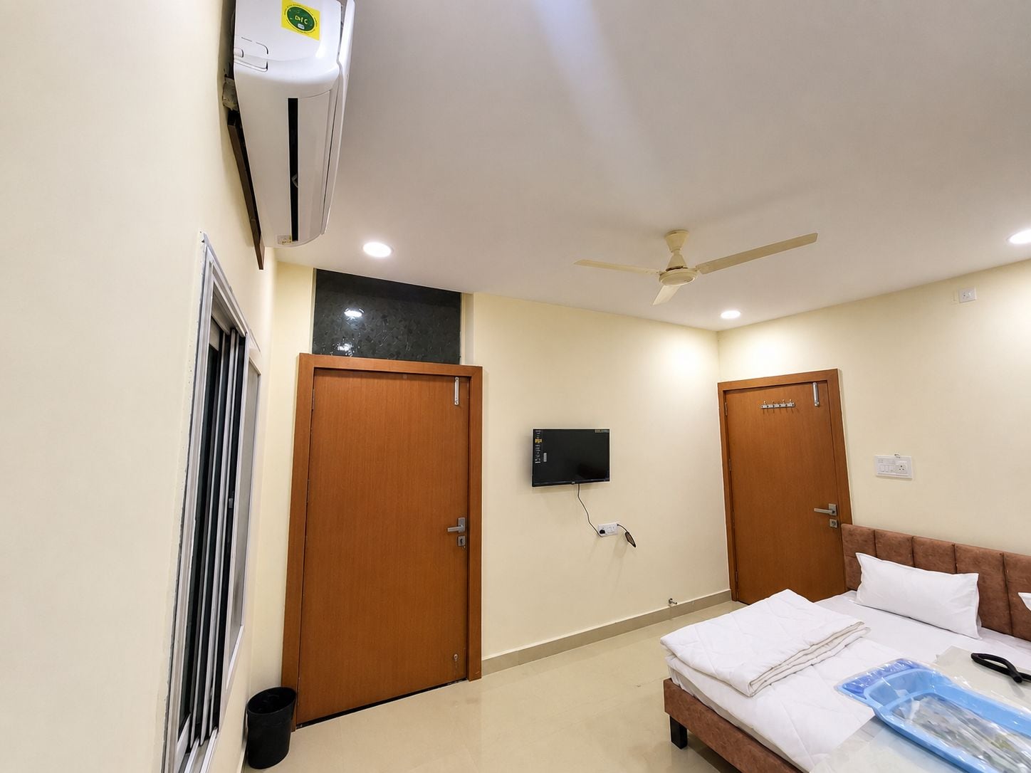 A hotel room with a bed, a ceiling fan, an air conditioning unit, and a television mounted on the wall.