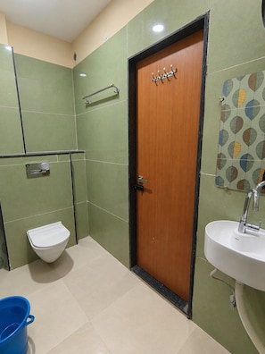 A bathroom with a wooden door, a white sink, a toilet, and a blue bucket. - HOTEL SHREE MANGLA (Ujjain)