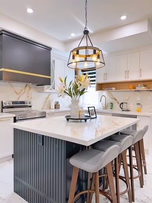 Uma cozinha moderna com uma ilha central, banquetas, forno de aço inoxidável e um lustre pendente.