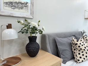 Une pièce avec un canapé gris, un vase noir contenant des fleurs blanches, une lampe de bureau blanche et un tableau encadré accroché au mur.