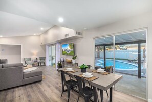 Ein modernes Wohnzimmer mit einem grauen Sofa, einem Essbereich mit Tisch und Stühlen, einem an der Wand befestigten Flachbildfernseher und einer großen Schiebeglasdoor zur Swimmingpool-Anlage.