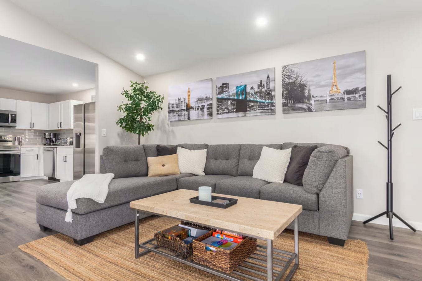 A modern living room with a grey sectional sofa, a coffee table, and a bookshelf. There are framed pictures on the wall, a plant, and a coat rack.