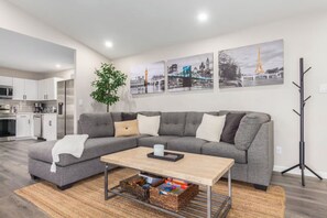 A modern living room with a grey sectional sofa, a coffee table, and a bookshelf. There are framed pictures on the wall, a plant, and a coat rack. - HEATED POOL - Minutes to Westgate District & Stadiums (Phoenix)