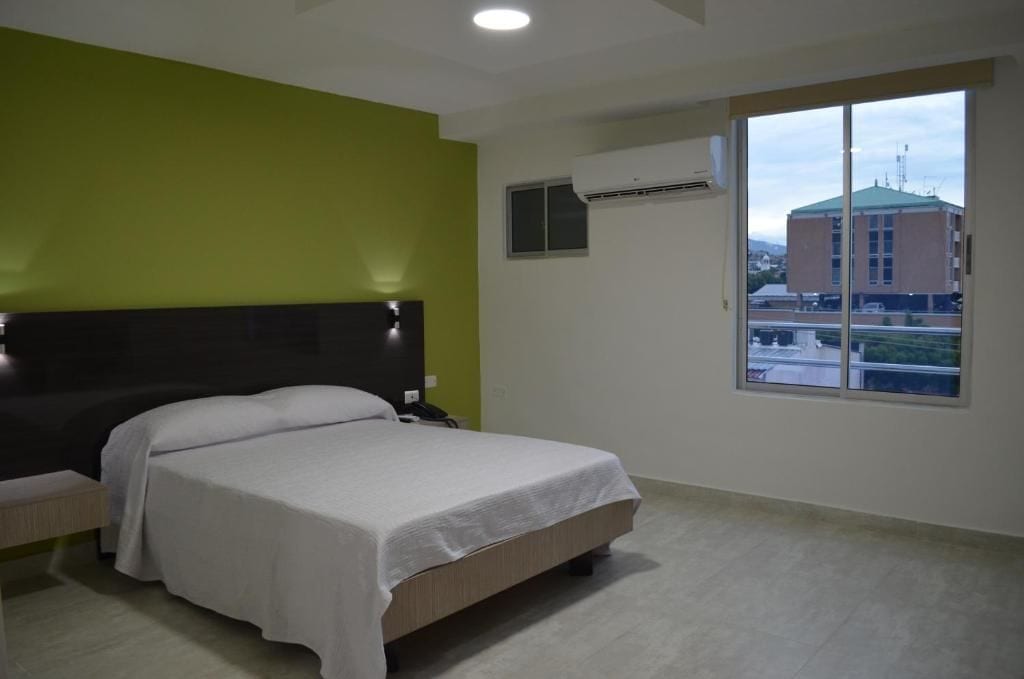 Habitación de hotel con cama, mesita de noche, equipo de aire acondicionado y ventana con vista a un edificio.