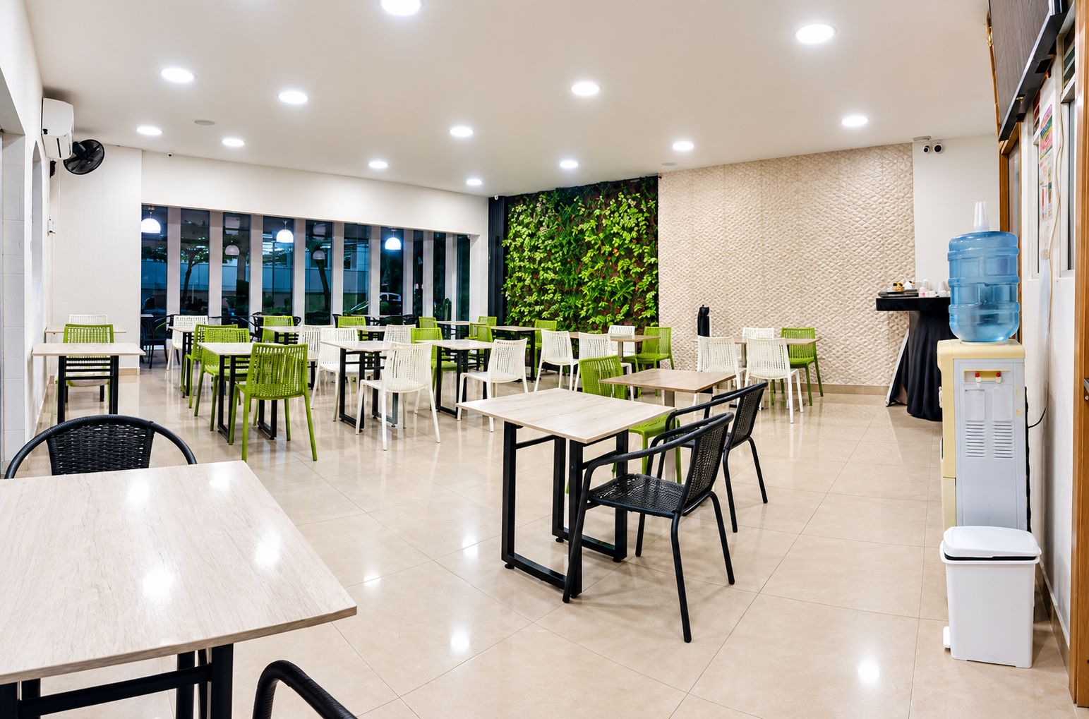 Uma área de refeições moderna com várias mesas e cadeiras, um dispensador de água e uma parede com uma planta verde.