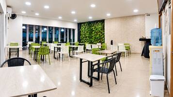 Uma área de refeições moderna com várias mesas e cadeiras, um dispensador de água e uma parede com uma planta verde.