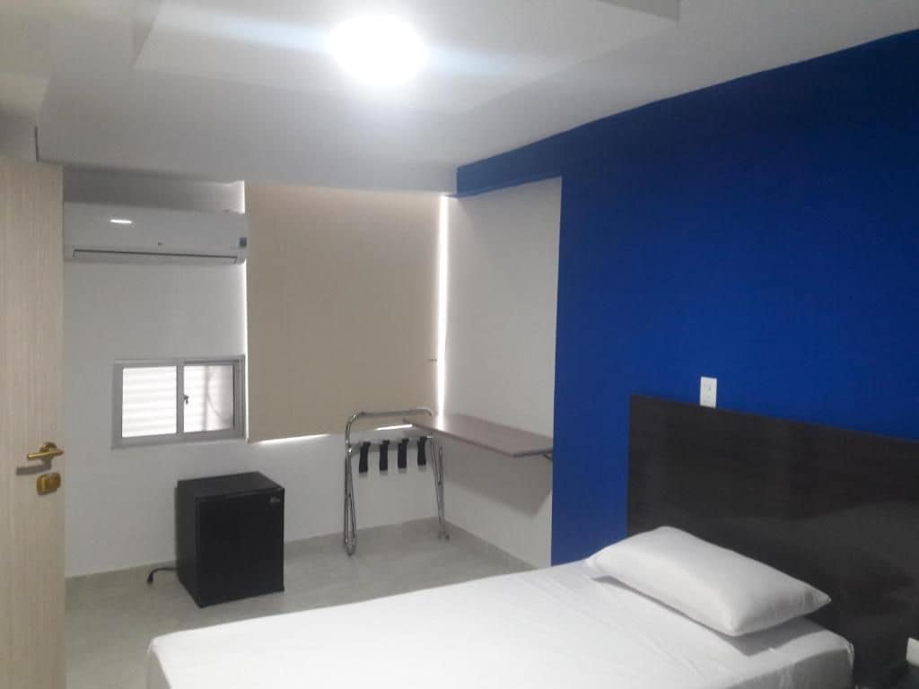 Un dormitorio con cama, una mesita de noche pequeña, ventana, calefactor y aire acondicionado instalado en la pared.