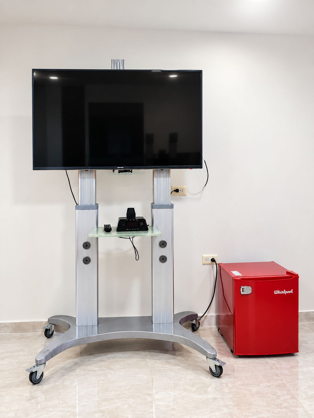 A television on a stand with wheels, a remote control, and a red appliance on the floor.