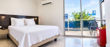 A hotel room with a bed, a bedside table with a phone, a red cabinet, and a balcony with a view of buildings and trees.