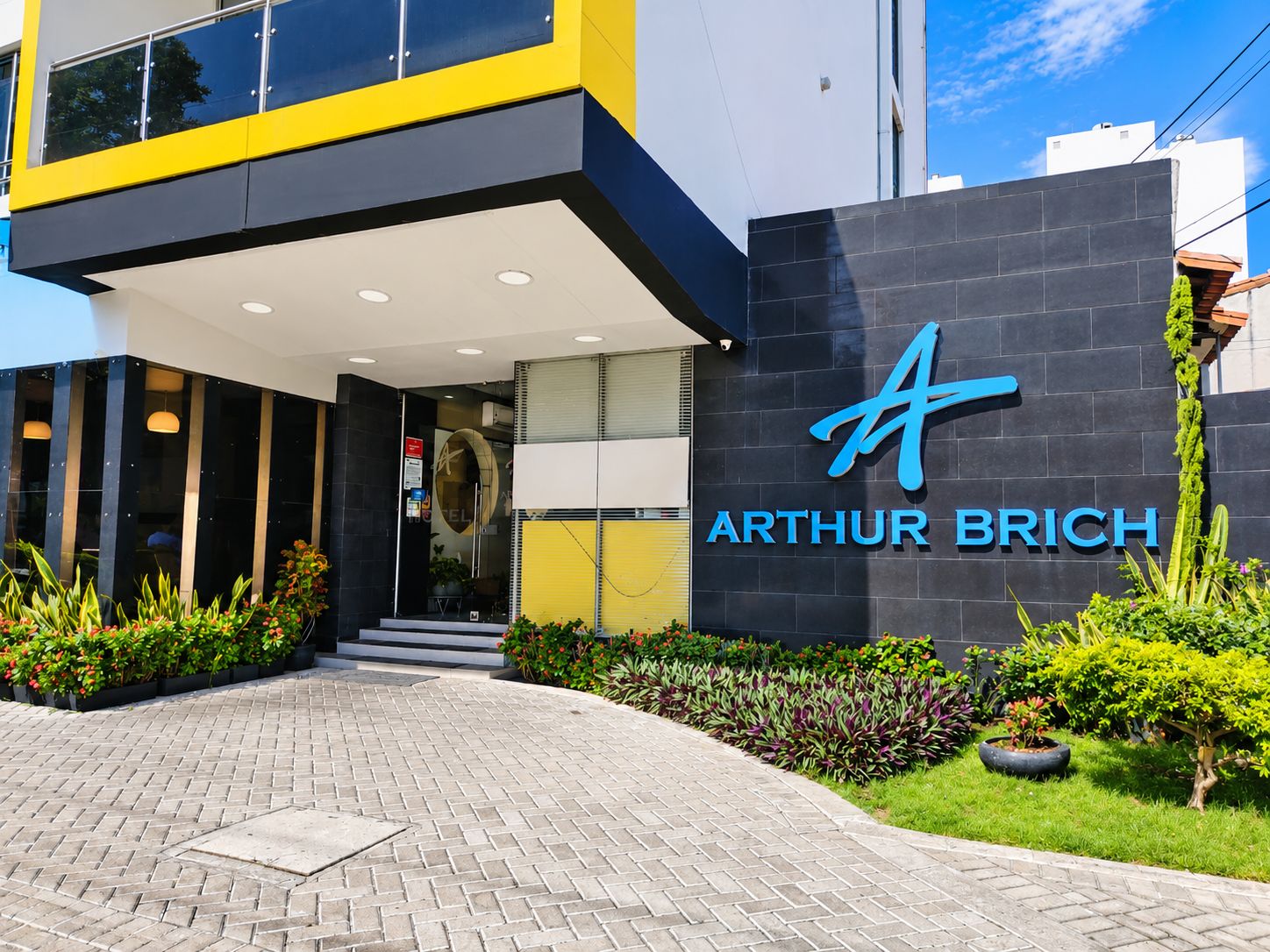 A modern hotel entrance with a prominent logo, a covered walkway, and well-maintained landscaping.