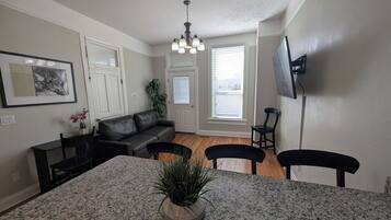 A living room with a black leather sofa, a dining table with chairs, a potted plant, and a ceiling light fixture.